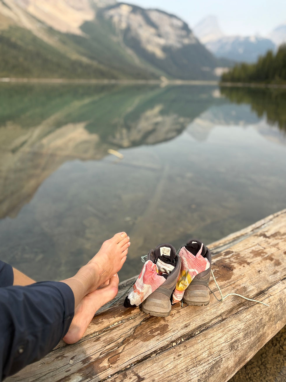 Excerpt from poem "Invest More" by Barefoot Theology displaying barefoot on a log with a mountains reflected in a still lake in the background