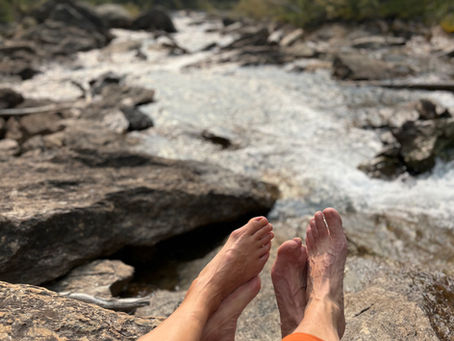 A man and woman's feet as they sit on a rock facing a mountain stream