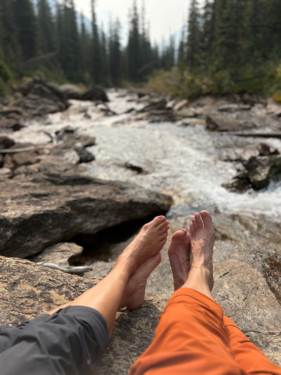A man and woman's feet as they sit on a rock facing a mountain stream