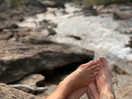 A man and woman's feet are touching and leaning into each other beside a mountain stream