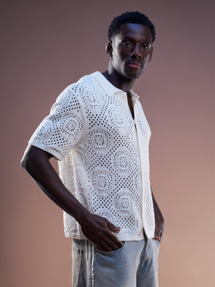 Man in witte gehaakte blouse met lichtgrijze / blauwe Adidas spijkerbroek voor een beige studioachtergrond. Het is een halfbody foto. 
