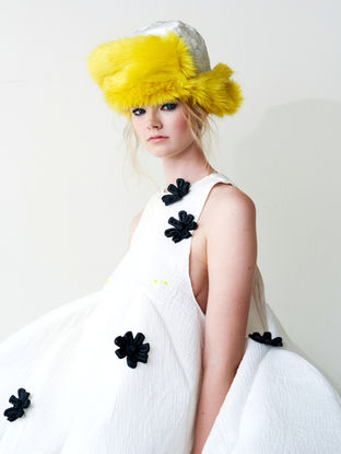Close up fashionportret van een model met blond haar, het model draagt een witte outfit voor een witte achtergrond. Het model draagt een hoed met gele details. 