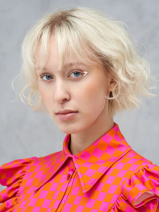 Close-up portret van een model met kort blond haar in een felroze en oranje geruite blouse. Witte achtergrond