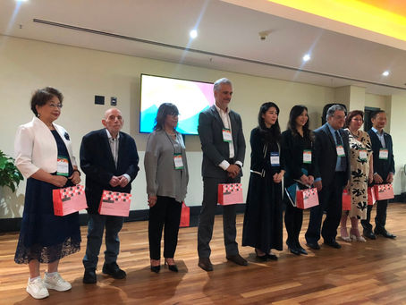 A imagem registra uma fila de nove pessoas posicionadas lado a lado sobre um piso de madeira em um ambiente de eventos formal, com crachás de identificação no peito e sacolas de presente estampadas em xadrez vermelho e branco nas mãos. Os participantes, dressed em trajes sociais como ternos, vestidos e blazers, parecem estar no palco de uma cerimônia corporativa ou conferência, com uma tela de exibição iluminada ao fundo e iluminação suave no teto.