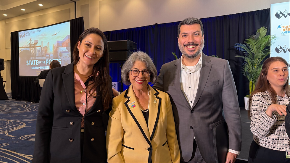 A foto registra um encontro institucional durante o evento "State of the Ports", realizado pelo World Trade Center Miami. Ao centro, destaca-se a Comissária (referida no contexto como Prefeita de distrito) Eileen Higgins, vestindo um blazer amarelo; à sua esquerda, está Nathalia Xavier, diretora da CISBRA, e à sua direita, Mario Scangarelli, Diretor Executivo da mesma entidade. O trio sorri para a câmera em um ambiente de conferência, com um telão ao fundo exibindo a temática portuária e logística do evento.