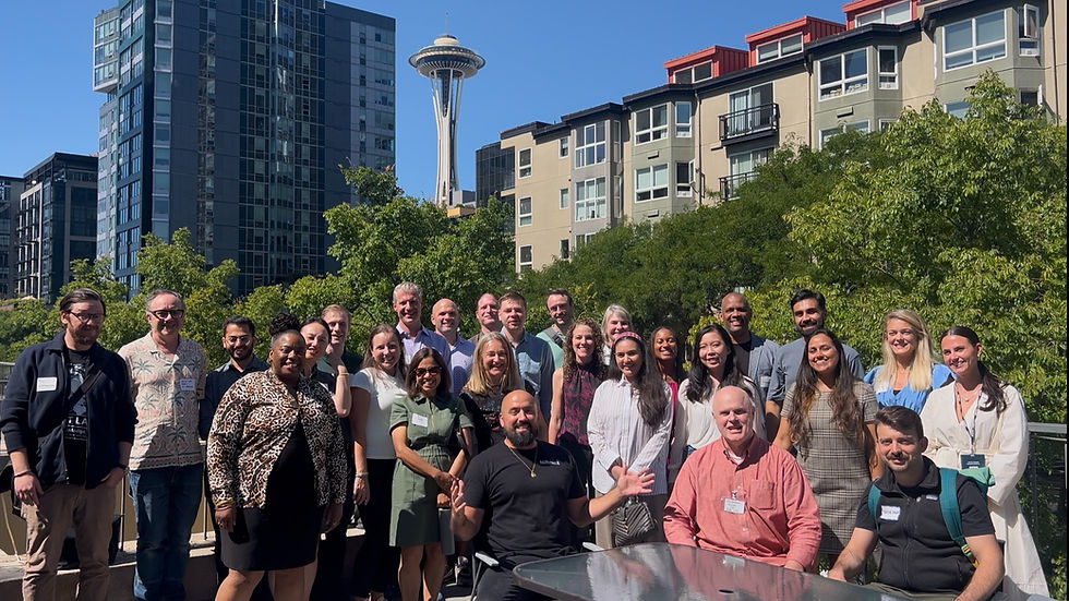 Group smiling on rooftop with Space Needle in background. Diverse people in casual wear. Sunny day, modern buildings, and greenery visible.