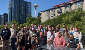 Group smiling on rooftop with Space Needle in background. Diverse people in casual wear. Sunny day, modern buildings, and greenery visible.
