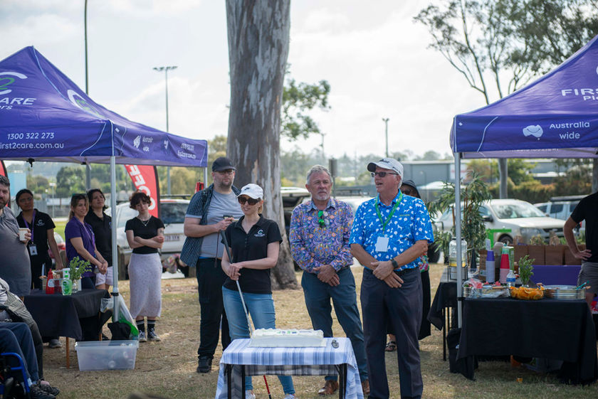 First2Care's annual Picnic in the park setup with tents, a group of people smiling and waiting for the cake to be cut.