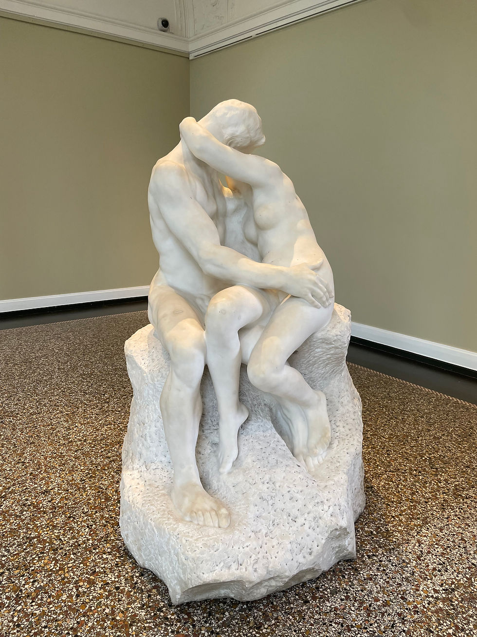 White sculpture of two nude figures embracing on a stone base in a museum room with green walls and speckled floor.
