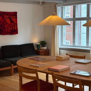 Warm dining room with wooden table, books, black sofa, red wall tapestry, and large windows under hanging cone lamps.