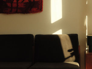 Sunlit living room with dark sofa, red wall hanging, green lamp on wooden table, and warm shadows across beige wall.