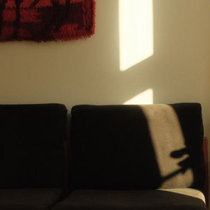 Sunlit living room with dark sofa, red wall hanging, green lamp on wooden table, and warm shadows across beige wall.
