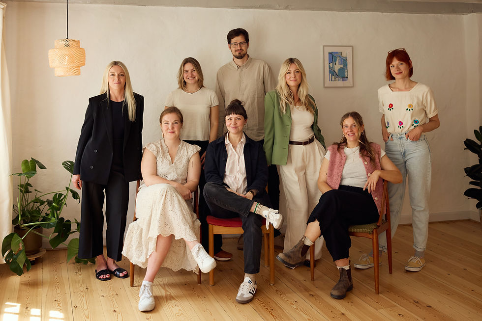 Seven smiling adults pose in a sunlit room with plants, wood floors, and a woven pendant lamp; one framed print hangs behind them.