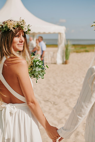 Braut mit Blumenschmuck im Haar läuft auf Pagodenzelt im Strand zu und guckt glücklich über ihre Schulter