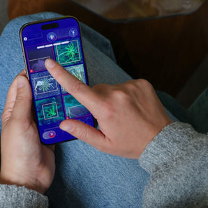 A person seated in a blue armchair plays a mini-game in the FathomVerse app on her iPhone. She is wearing a gray sweater and blue jeans. On her phone screen is a grid of six photos on a blue background with app icons. She is tapping one of the photos to add a red box. In the background is gray carpet.
