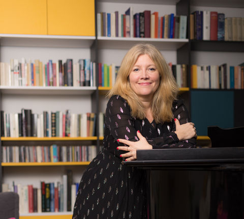 Sopranistin, Musikjournalistin und Moderatorin Eva Schramm – Portraitfoto in schwarzem Kleid vor Bücherregal auf einen Flügel gestützt.