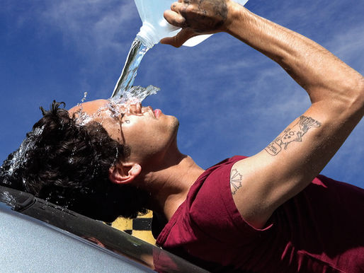 Dominic Fike laying down pouring water on his face wearing a maroon T-shirt with two tattoos on his right arm.