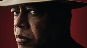 Blues artist Billy Branch on a red background wearing a hat looking into the camera.