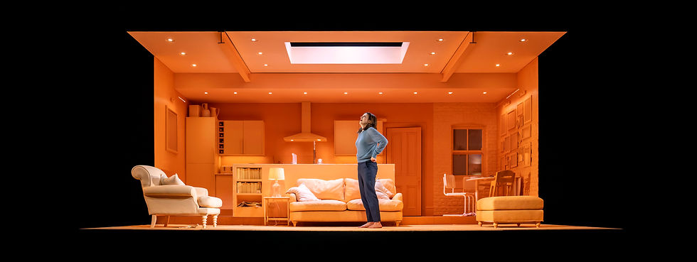 A woman, Aisling Loftus, with shoulder-length brown hair and wearing a blue jumper and blue trousers, stands in front of a sofa in a set of an apartment, where all the furniture is peach. She has her hands on her hips and is looking upwards, as if breathing a sigh of relief.