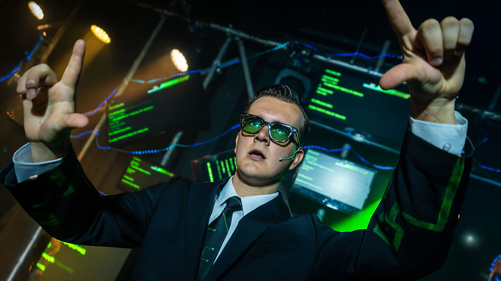 Man in a suit with green glasses and headset gestures dramatically. Background displays green text on screens, with stage lights overhead.
