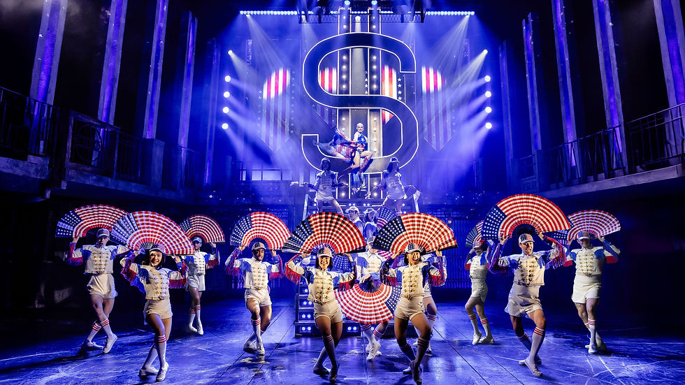 Performers in uniforms with red, white, and blue striped fans dance on stage, backlit by a large dollar sign and blue lights. Mood is energetic.