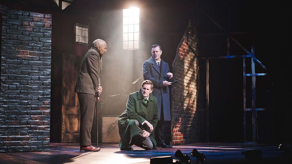 Three men in suits in a dimly lit brick-walled room. One kneels, another stands with a cane, and the third gestures with a clipboard. Dramatic mood.