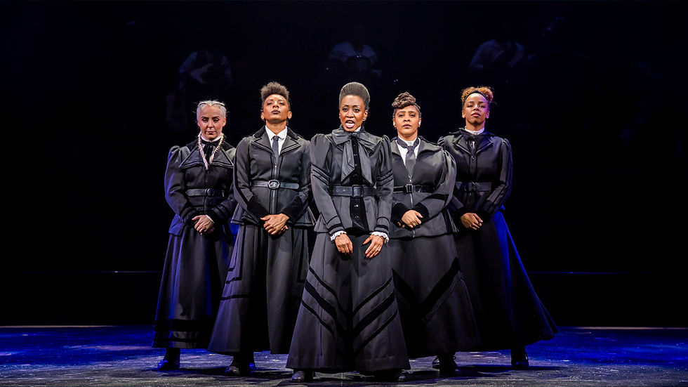 Five women in black period costumes stand on a dark stage under blue light, facing forward with stern expressions.