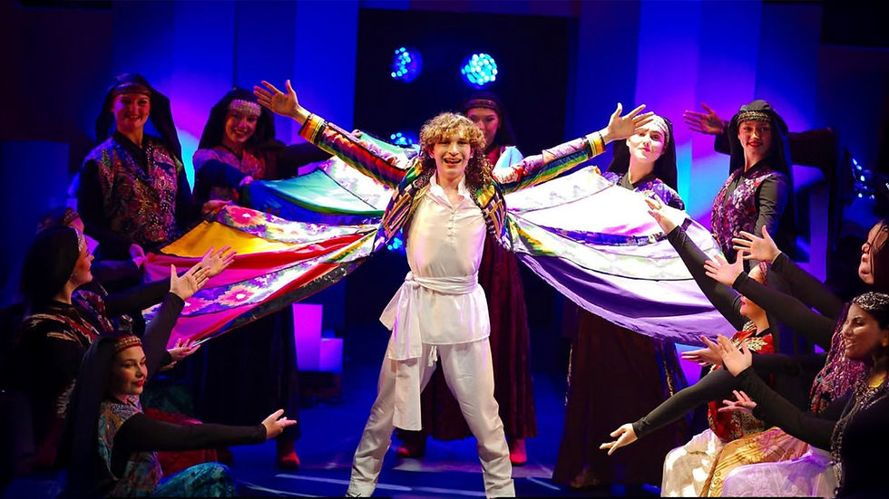 Performer in a colorful coat with outstretched arms onstage, surrounded by dancers in vibrant attire, under purple lights. Joyful mood.