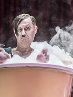 Man with stern expression sits in a frothy bathtub, surrounded by steam. Red curtain backdrop enhances theatrical mood.