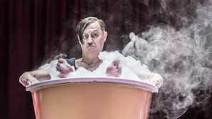 Man with stern expression sits in a frothy bathtub, surrounded by steam. Red curtain backdrop enhances theatrical mood.