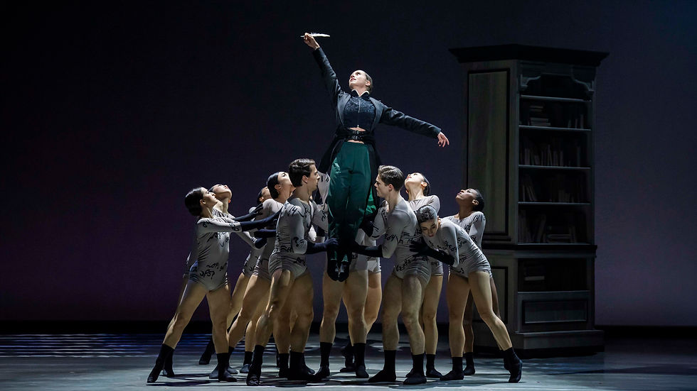 Dancers in gray outfits lift a person in dark clothing reaching upwards, on a stage with a bookshelf background. The scene is dramatic and expressive.
