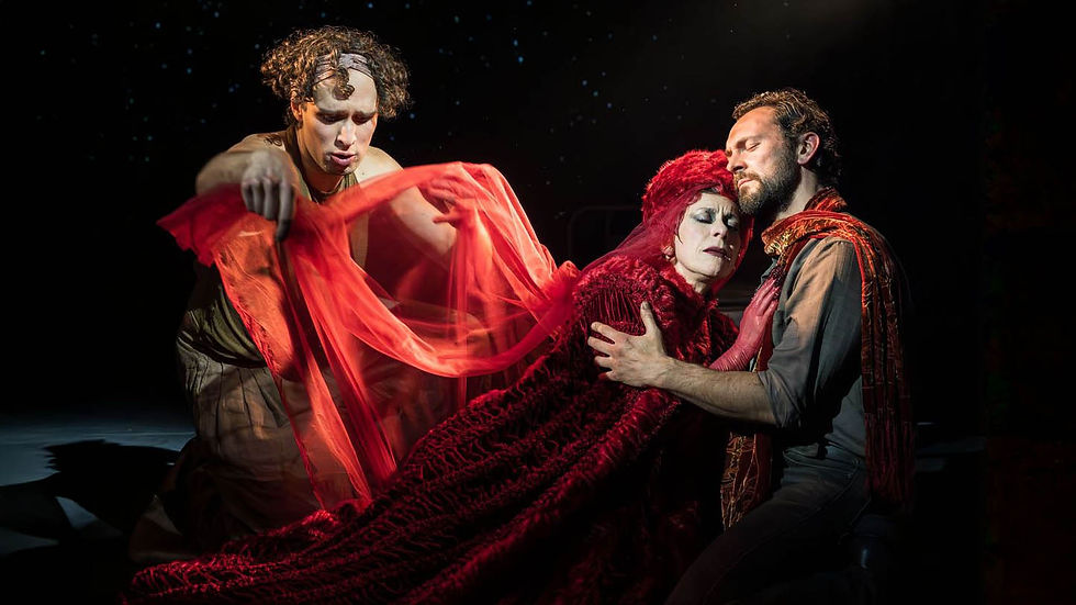 Three people performing on stage. One holds a red cloth dramatically, while two embrace. Dark background enhances theatrical mood.