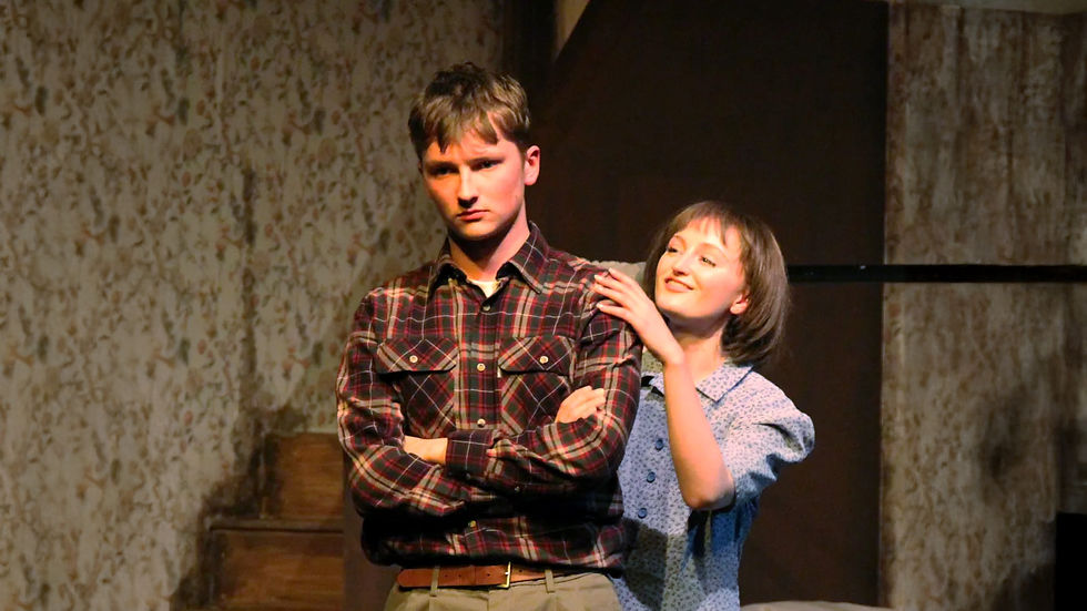 A woman gently touches a gay man's shoulders; he's in a plaid shirt with arms crossed, looking serious. They're indoors against a patterned wall.