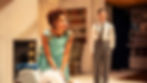 Woman in teal dress leans on a chair, looking concerned. Man in suit stands in background. Room with bookshelf and fireplace. Vintage setting.