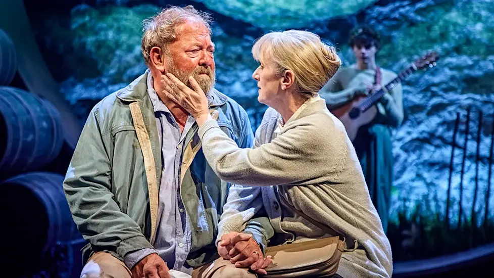 An elderly woman gently holds a man's face on stage, creating an emotional moment. A guitarist stands in the background. Blue-toned backdrop.
