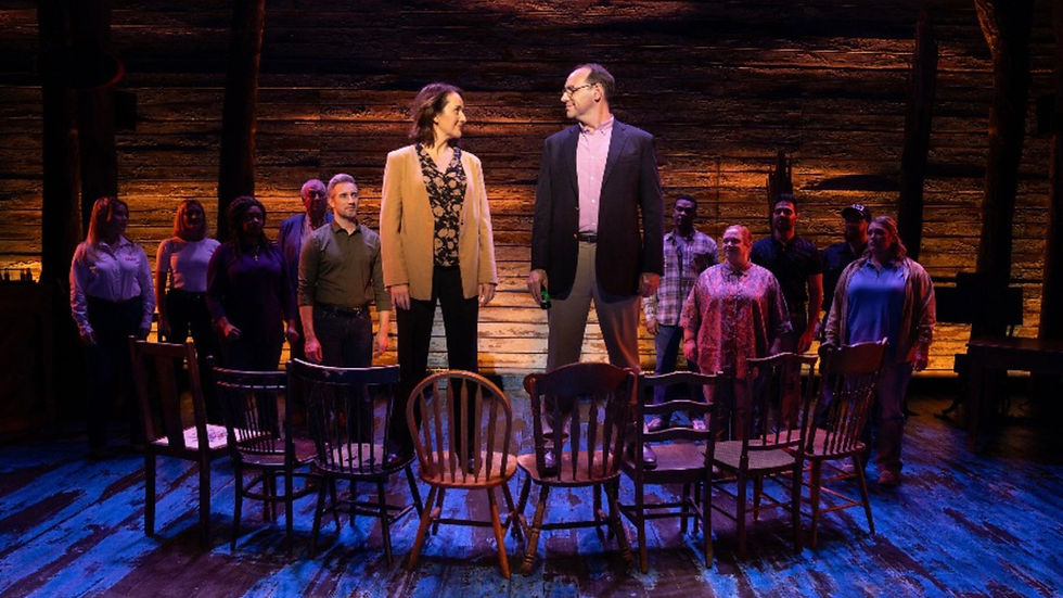 A man and woman both in jackets standing on a line of wooden chairs on a stage which is lit blue and orange, from the musical Come From Away