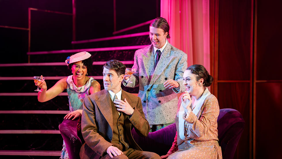 Four people in vintage attire laugh on a stage set with pink lighting. Two hold drinks while the seated man appears surprised.