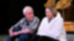 Things I Know To Be True Play. An older man in a plaid shirt sits beside a concerned woman wrapped in a white blanket. They are on stage with golden trees in the background.