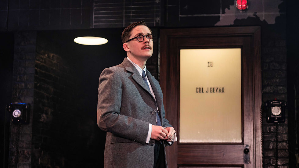 Man in a gray suit and glasses stands on a dim stage before a door labeled Col J Beval, looking up thoughtfully.