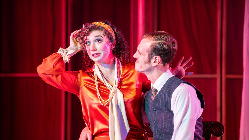 A woman in an orange dress and pearls speaks animatedly next to a man in a suit. Red curtains in the background set a dramatic mood.