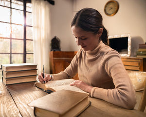 Woman writing in a book at a wooden table, surrounded by stacked books. Sunlight streams through a window, creating a warm, focused mood.