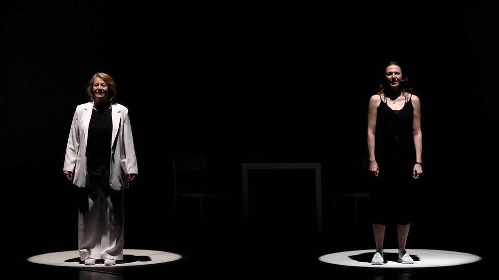 Two women stand in spotlights on a dark stage, one in a white suit and one in a black dress, with chairs and a table behind them.