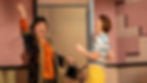 Two women joyfully interacting indoors. One wears vibrant orange and black, the other in blue stripes and yellow. Pink patterned wall backdrop.