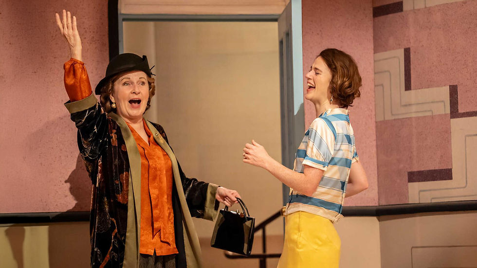 Two women joyfully interacting indoors. One wears vibrant orange and black, the other in blue stripes and yellow. Pink patterned wall backdrop.