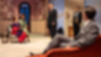 Five people in formal attire are in a stylish room with geometric patterns. Two women sit, two men stand, and one man reclines on a chair.