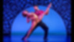 Two ballroom dancers pose on a blue-lit stage, man supporting a woman in a dramatic leg lift against a black arch backdrop.