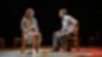 Two actors sit on wooden chairs on a dark theater stage, facing each other in tense conversation under warm spotlight.