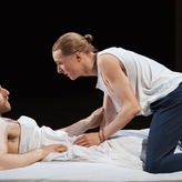Two people in casual clothes on a bed, one lying down and one kneeling, engaged in an intense conversation. Dimly lit setting.