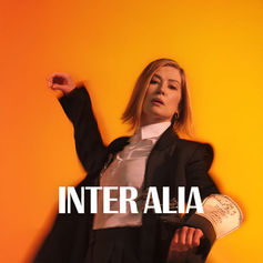 Person in a suit with one arm raised, set against an orange background. "INTER ALIA" text is prominent. Mood is confident and stylish.