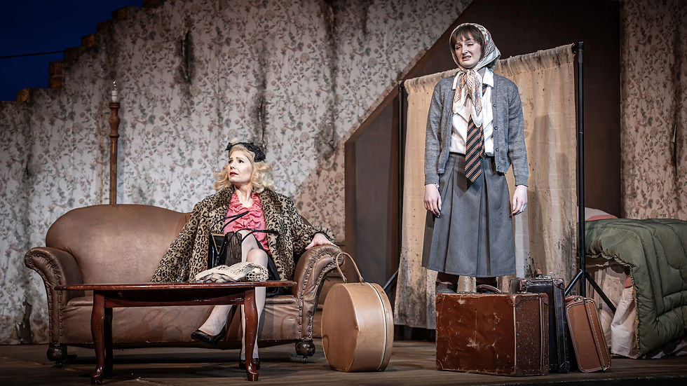 Two women in vintage attire on stage; one seated on a sofa with a leopard coat, the other standing by luggage. Floral wallpaper background.